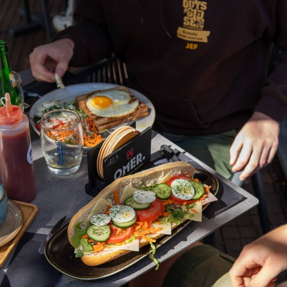 Een belegde baguette met kaas, hesp, tomaat, komkommer en ei, en een croque met spiegelei op de achtergrond.