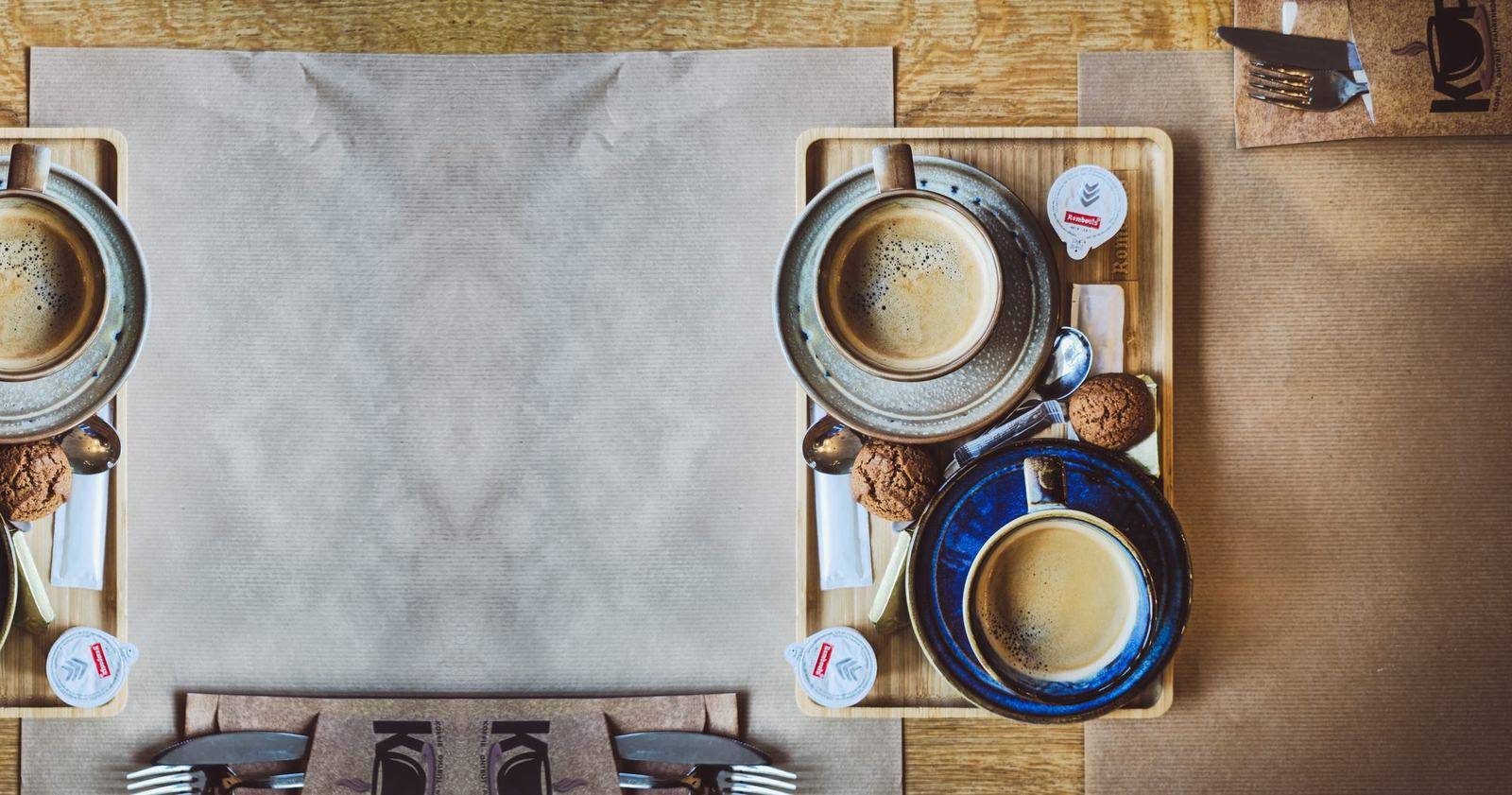 Twee koffies op een houten bamboe dienblad, met speculoos, melk en suiker, op een tafel met bruin papier en KOP-bestek.