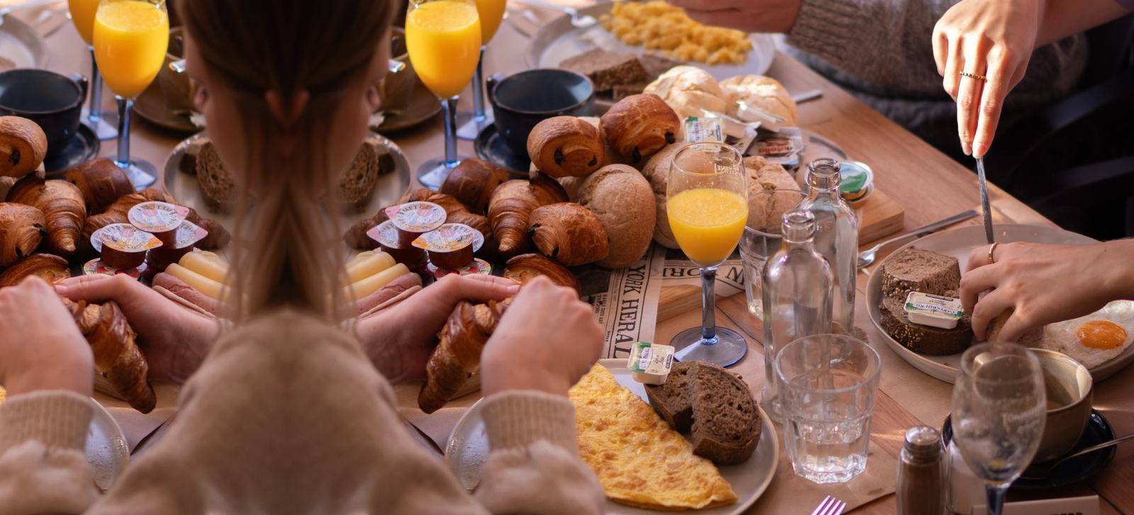 Een gezelschap deelt een ontbijtplank met croissants, broodjes, kaas en hesp, met verse sinaasappelsap en bubbels.