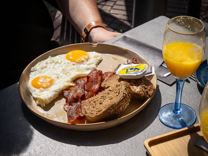 Twee spiegeleieren met krokant spek, bruin brood en een glas vers sinaasappelsap in de zon.
